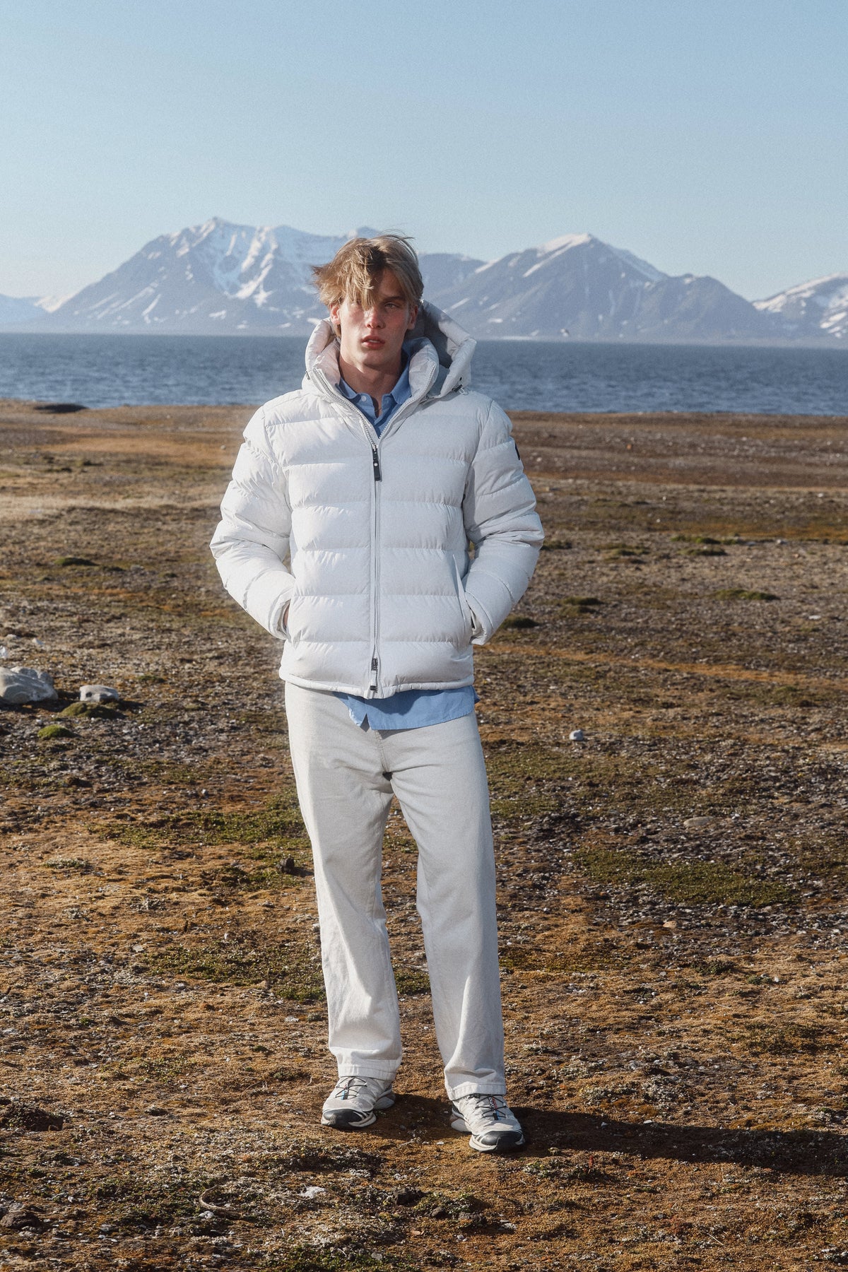 A man wearing a white puffer jacket, light beige trousers, and a light blue shirt stands on a rocky, grassy shoreline with water and snow-capped mountains in the background.